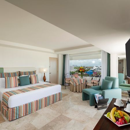 Отель Grand Oasis Palm Канкун Экстерьер фото The photo shows a spacious hotel room with modern decor. It features a large bed with a striped duvet and decorative pillows, flanked by two lamps. There are comfortable seating areas with light green chairs and a small table. A balcony can be seen i
