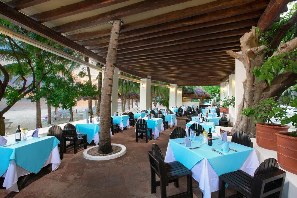 Отель Grand Oasis Palm Канкун Экстерьер фото The photo shows an outdoor dining area of a restaurant. The space features several tables covered with light blue tablecloths, each accompanied by black wooden chairs. The area is shaded by a wooden roof supported by columns, and there are leafy tree