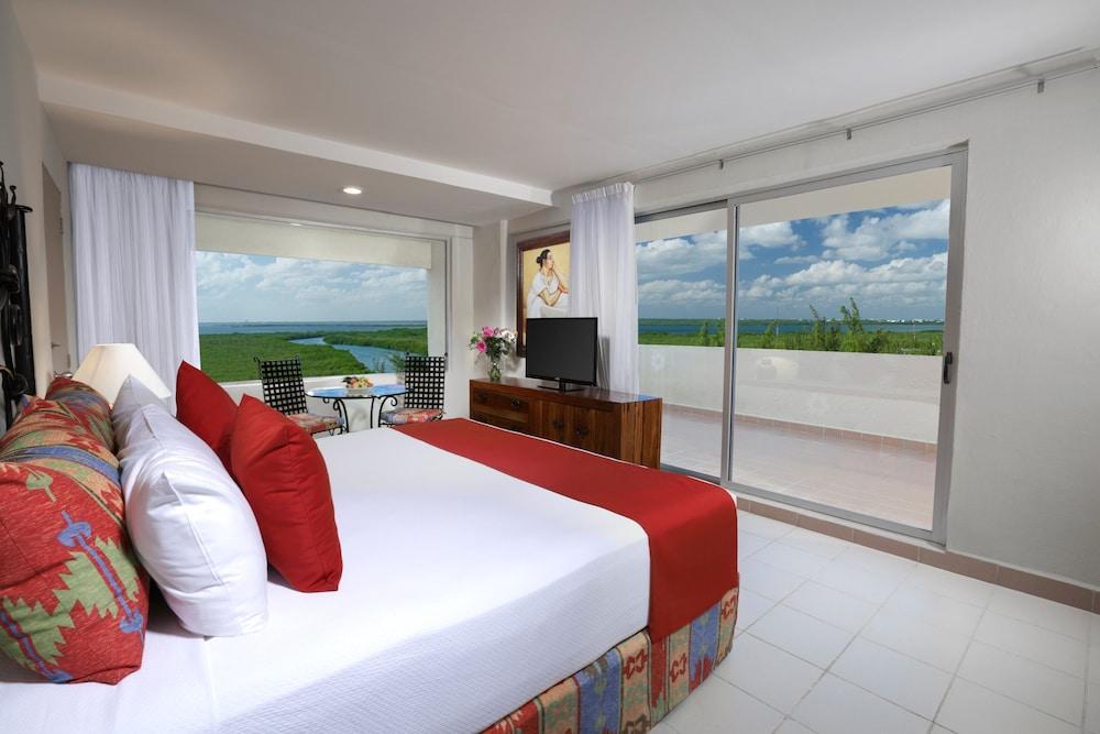 Отель Grand Oasis Palm Канкун Экстерьер фото The photo shows a modern hotel room featuring a large bed with white linens and red accents. There are decorative pillows on the bed, and a bedside table with a lamp. To the side, there’s a large window with white curtains that offers a view of a bal