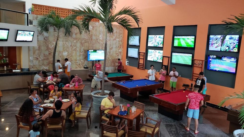 Отель Grand Oasis Palm Канкун Экстерьер фото The photo shows a lively recreational area inside a building, likely a bar or game room. There are several pool tables, one blue and one red, where people are playing or waiting for their turn. 

Around the area, groups of people are seated at tables