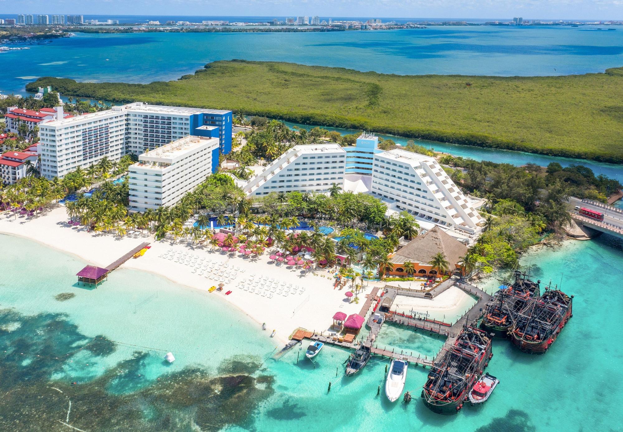 Отель Grand Oasis Palm Канкун Экстерьер фото The photo shows a beautiful coastal resort area. In the foreground, there is a sandy beach with lounge chairs and umbrellas, while the clear turquoise waters are visible. The resort features large white and blue buildings, with lush greenery surround