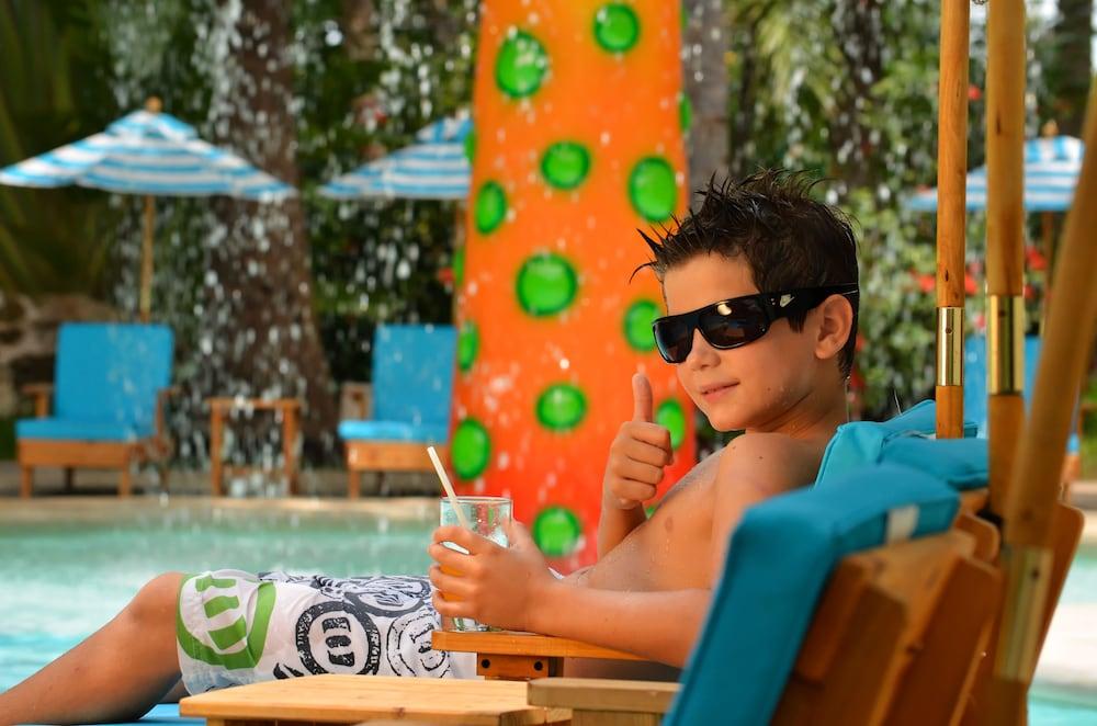 Отель Grand Oasis Palm Канкун Экстерьер фото The photo shows a young boy relaxing by a pool. He is wearing sunglasses and has a thumbs-up gesture, signaling enjoyment. He appears to be lounging on a chair, holding a drink in a glass. In the background, there is a bright and colorful structure w