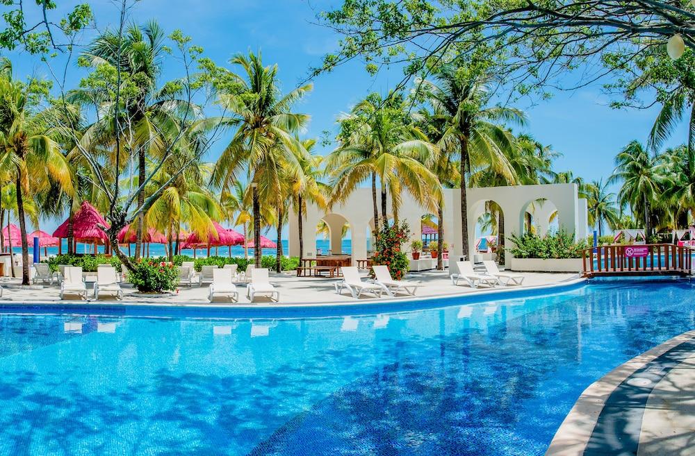 Отель Grand Oasis Palm Канкун Экстерьер фото The photo depicts a tropical resort scene featuring a large, inviting swimming pool surrounded by palm trees. The water in the pool is bright blue, reflecting the clear sky above. There are several white lounge chairs arranged around the pool area, p