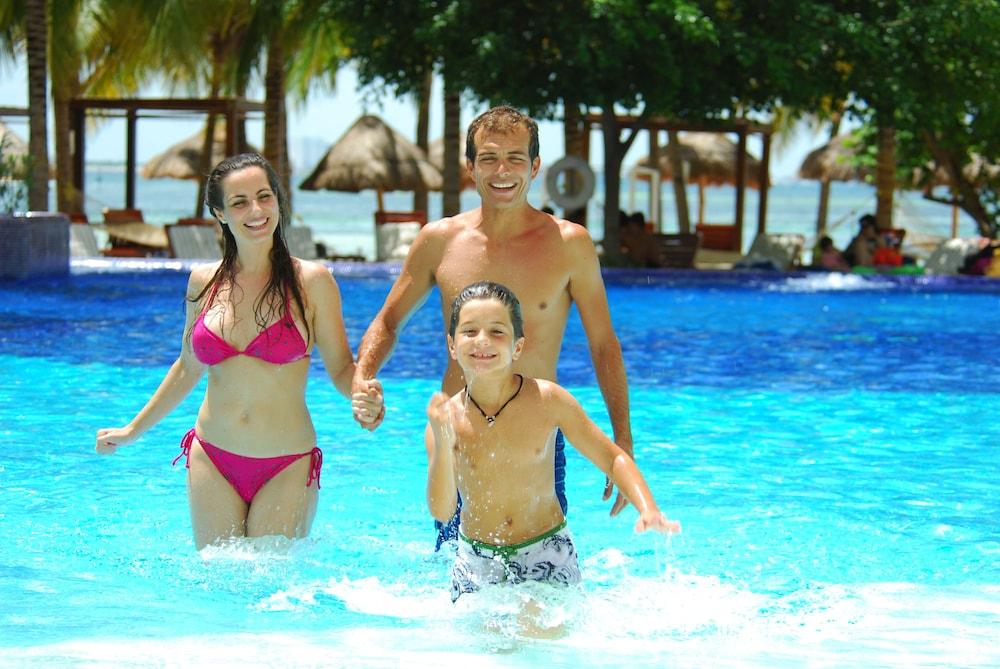 Отель Grand Oasis Palm Канкун Экстерьер фото The photo depicts a family enjoying a day at a pool. A woman in a pink bikini and a man are holding hands, while a boy runs happily in the water beside them. The pool is bright blue, and the background features palm trees and thatched huts, suggestin