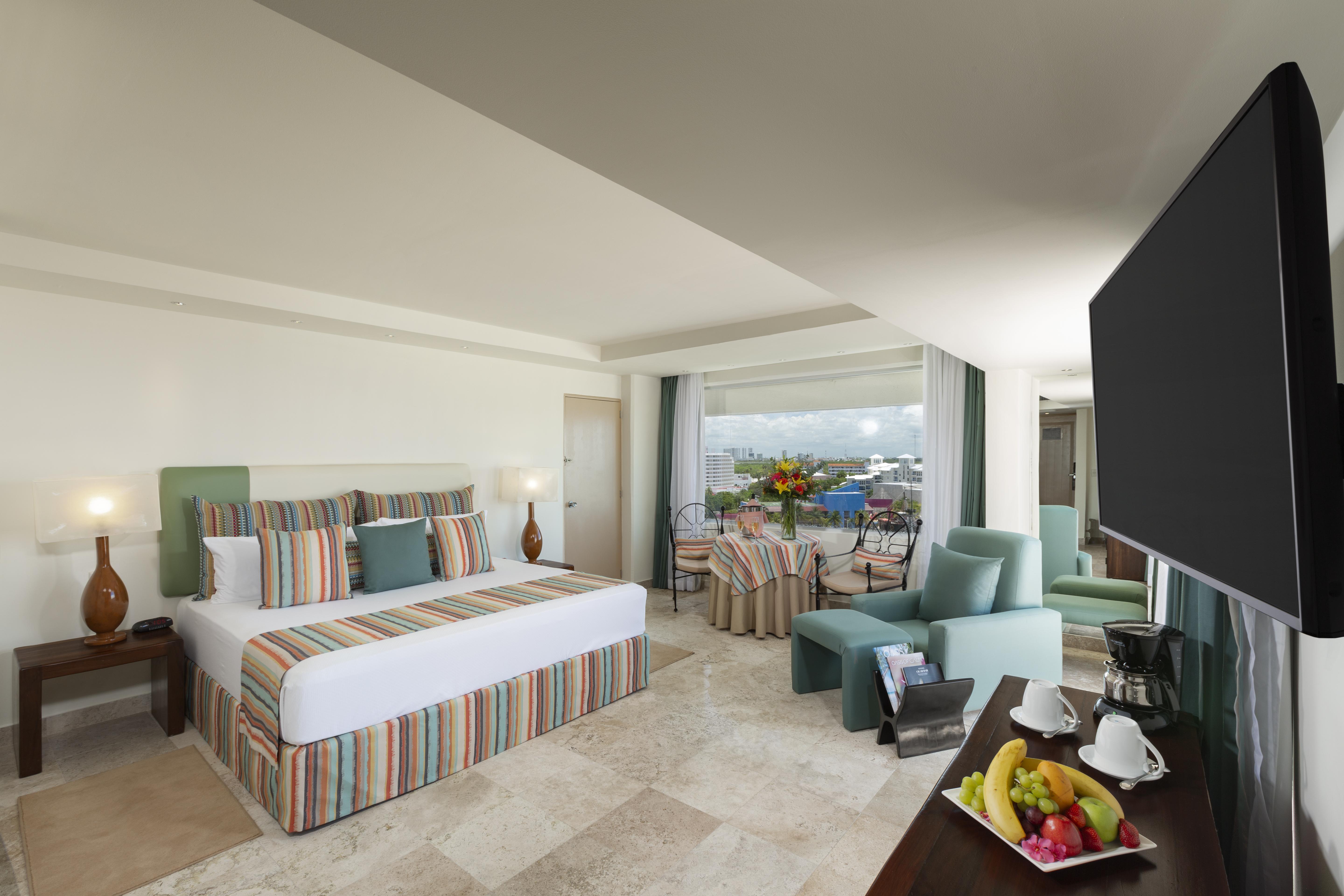 Отель Grand Oasis Palm Канкун Экстерьер фото The photo shows a spacious hotel room with modern decor. It features a large bed with a striped duvet and decorative pillows, flanked by two lamps. There are comfortable seating areas with light green chairs and a small table. A balcony can be seen i