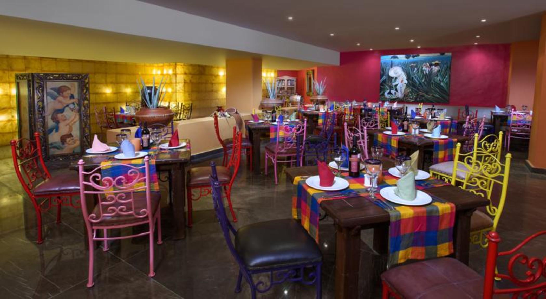 Отель Grand Oasis Palm Канкун Экстерьер фото The photo shows a vibrant and colorful restaurant interior. There are several tables arranged with multicolored tablecloths, each adorned with plates and decorative napkins. The chairs are also colorful, featuring a mix of pink, purple, and metal des