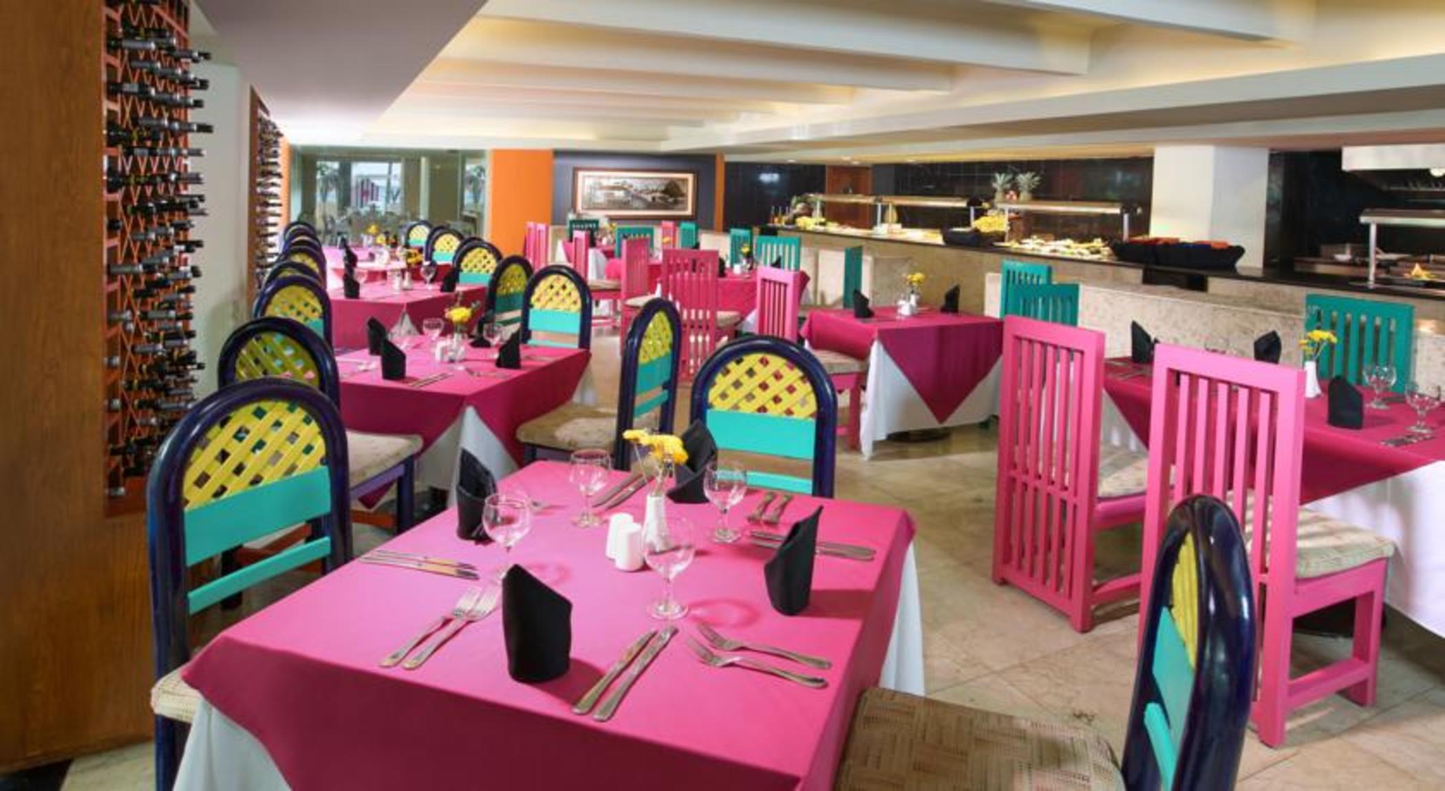 Отель Grand Oasis Palm Канкун Экстерьер фото The photo shows a vibrant restaurant interior featuring colorful decor. There are multiple tables covered with bright pink tablecloths. Each table is equipped with utensils, napkins, and a small centerpiece, possibly a flower. The chairs are painted 