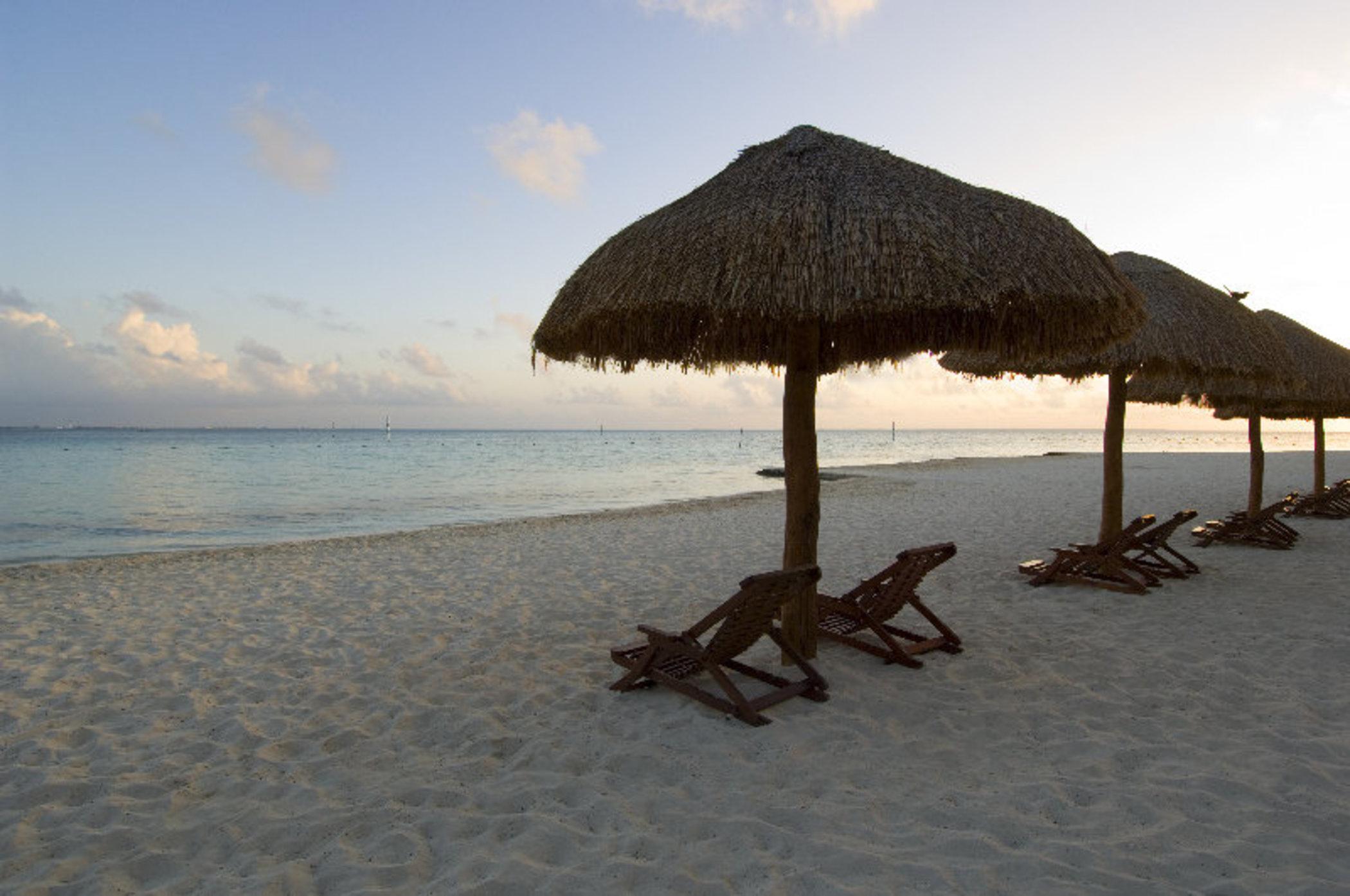 Отель Grand Oasis Palm Канкун Экстерьер фото The photo depicts a serene beach scene featuring several thatched umbrellas on the sand. Beneath the umbrellas are wooden lounge chairs, positioned for relaxation. The beach has soft, white sand, and the calm waters of the sea reflect the colors of t