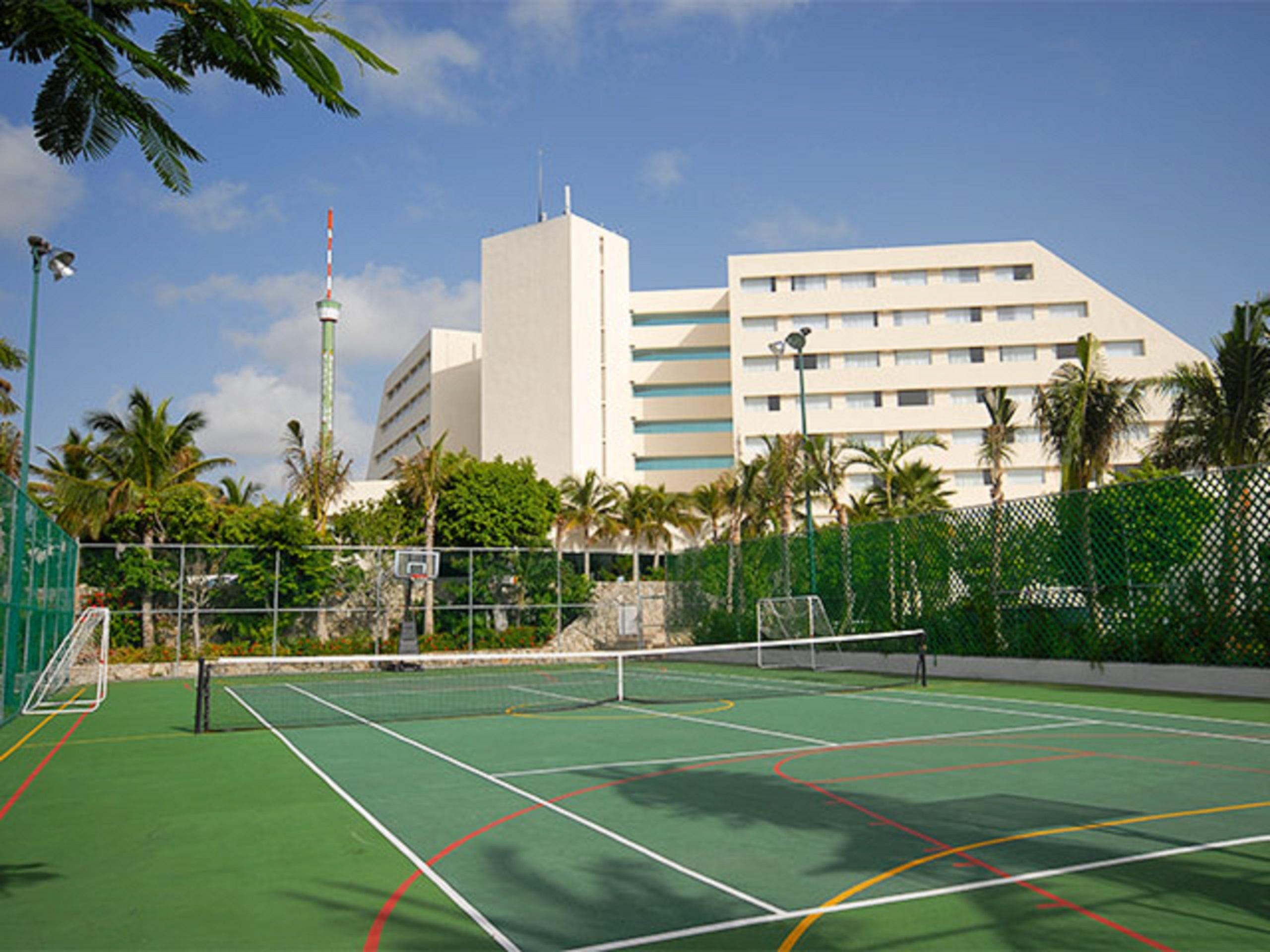 Отель Grand Oasis Palm Канкун Экстерьер фото The photo shows a sports facility featuring a tennis court in the foreground. The court is surrounded by a green fence and has distinctive lines marking the playing area. In the background, there is a tall, modern building, possibly a hotel or resort