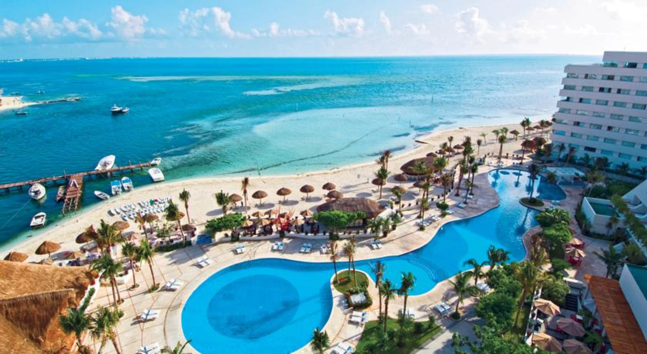 Отель Grand Oasis Palm Канкун Экстерьер фото The photo depicts a beautiful resort area located near a beach. In the foreground, there are two large swimming pools, one of which features a curved shape, surrounded by lounge chairs. Palm trees and tropical plants are scattered throughout the area