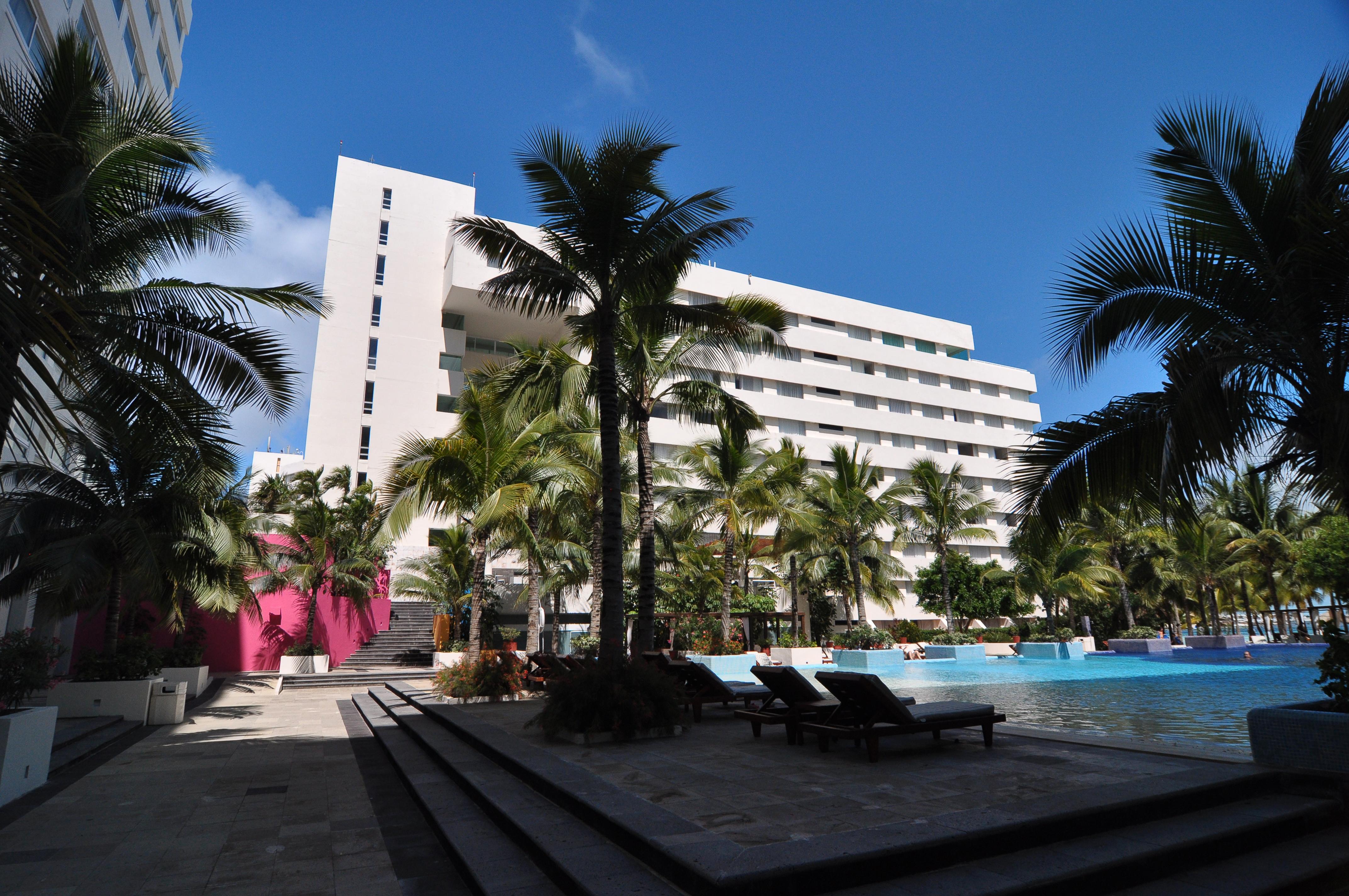Отель Grand Oasis Palm Канкун Экстерьер фото The photo showcases a modern building, likely a hotel or resort, situated next to a swimming pool. The structure is white and has multiple levels, surrounded by lush palm trees, giving a tropical feel. There is a prominent pink wall in the background