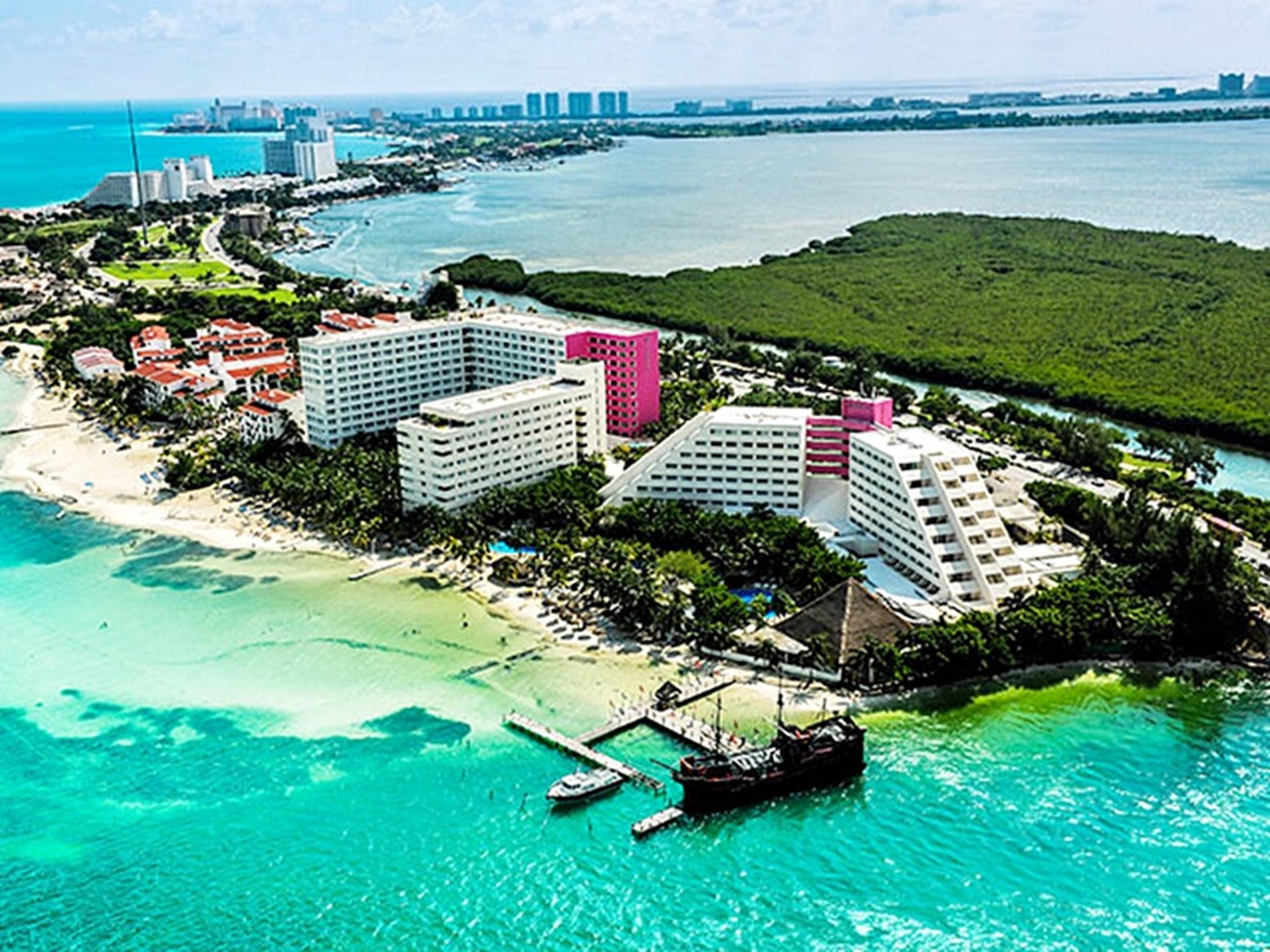 Отель Grand Oasis Palm Канкун Экстерьер фото The photo depicts an aerial view of a coastal area, showcasing several buildings along the shoreline. The structures include a mix of white and pink hotels or resorts, surrounded by lush greenery and sandy beaches. The water is a vibrant turquoise co