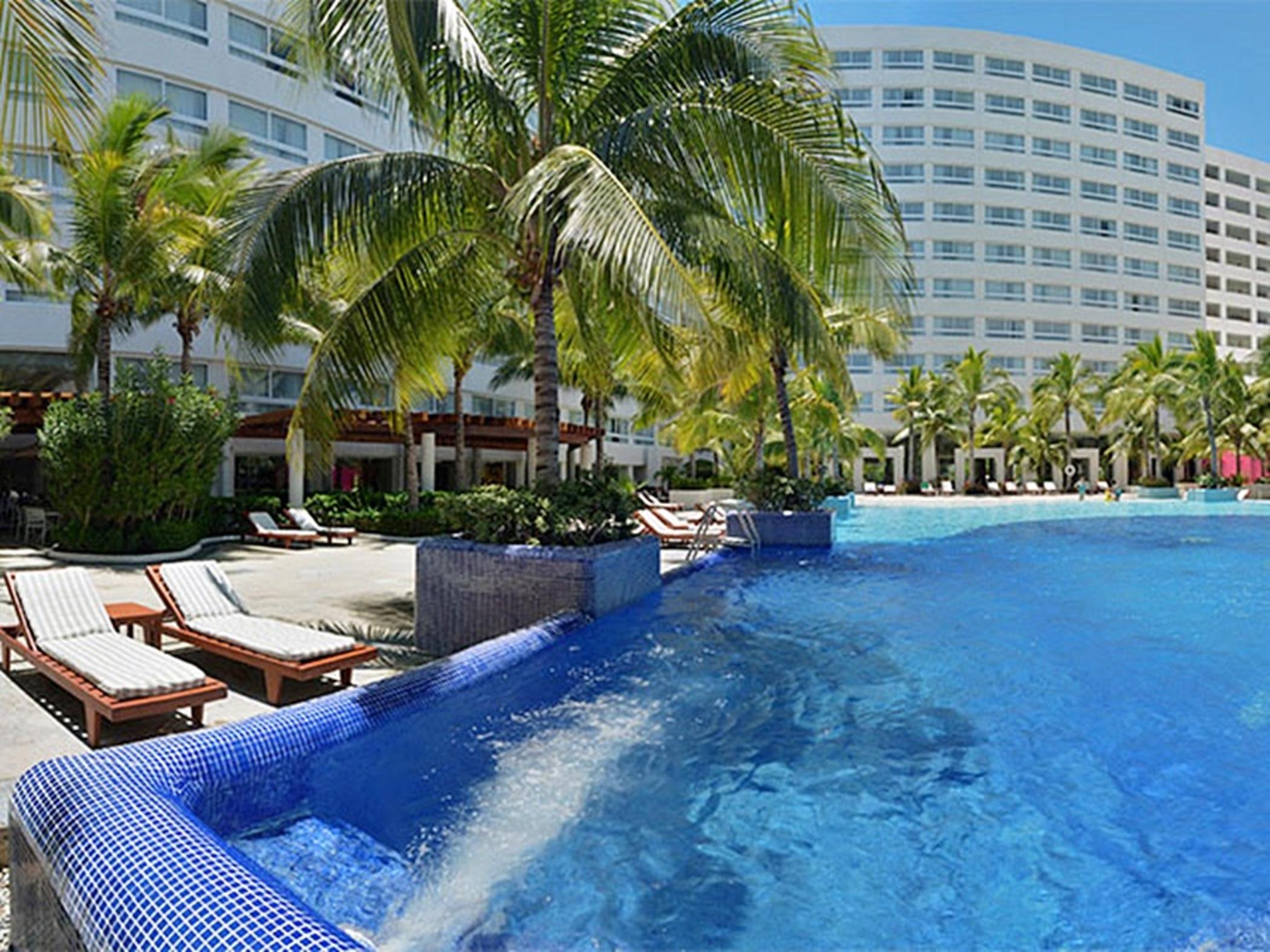 Отель Grand Oasis Palm Канкун Экстерьер фото The photo shows a resort-style setting featuring a large swimming pool with clear blue water. Surrounding the pool are lounge chairs, some of which are positioned under palm trees, providing a tropical ambiance. In the background, there is a multi-st