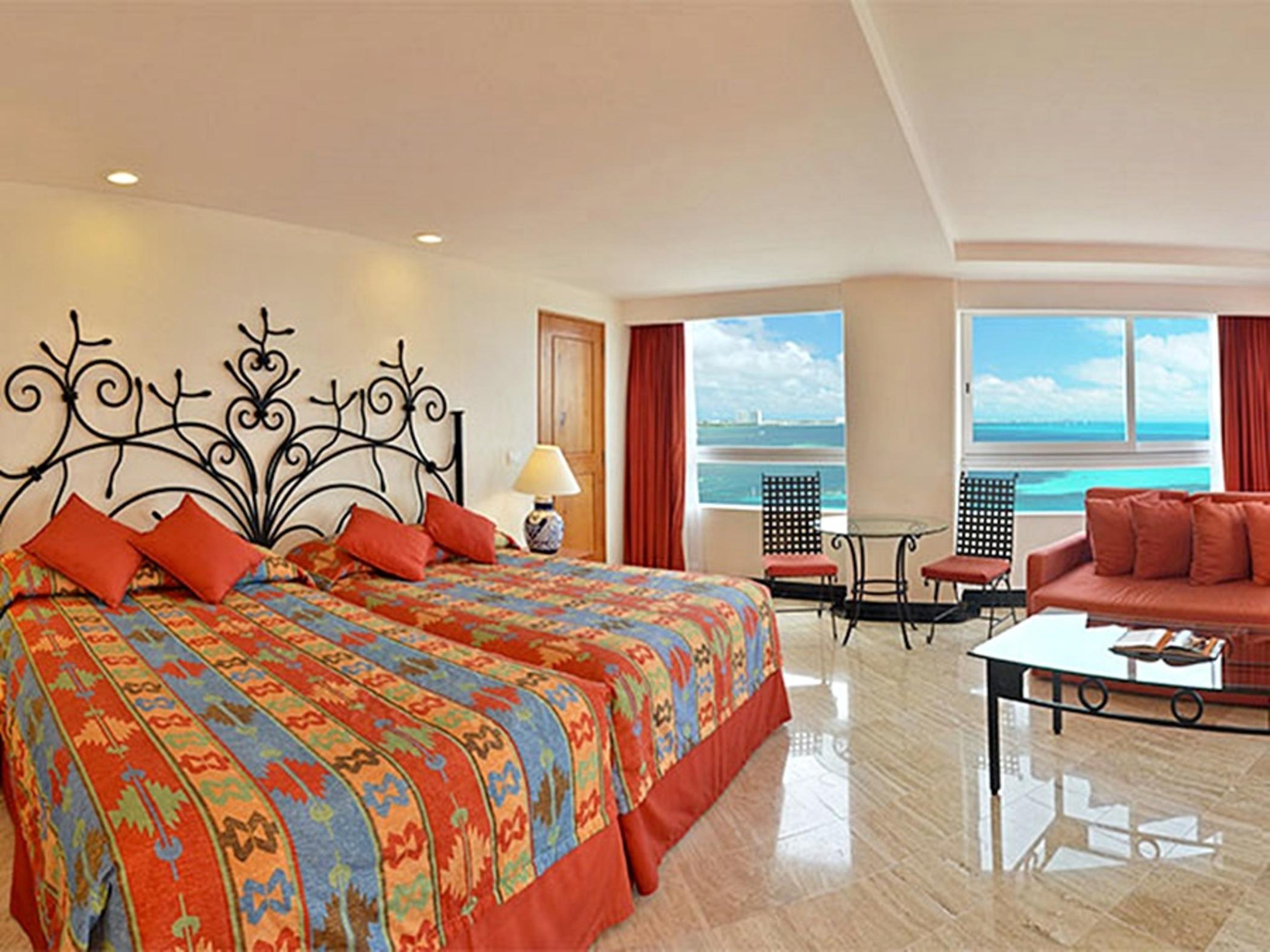 Отель Grand Oasis Palm Канкун Экстерьер фото The photo shows a spacious hotel room featuring two beds with colorful patterned bedding. The walls are decorated with a decorative iron headboard. There are large windows that provide a view of the ocean and a bright, sunny sky. The room is warmly l