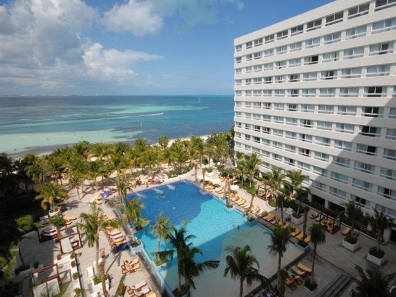 Отель Grand Oasis Palm Канкун Удобства фото The image shows a luxurious hotel resort situated by the ocean. In the foreground, there is a large swimming pool surrounded by palm trees and sun loungers, creating a relaxing atmosphere for guests. Beyond the pool, the beach and ocean can be seen, 