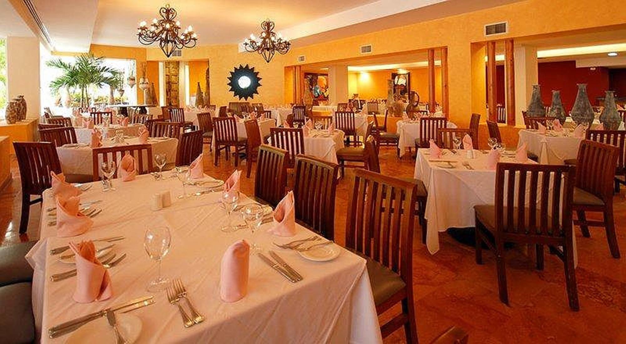 Отель Grand Oasis Palm Канкун Ресторан фото The photo shows an interior of a restaurant with an inviting and elegant ambiance. There are several tables set up, each covered with white tablecloths. The place settings include glassware, silverware, and neatly folded pink napkins. The walls are p