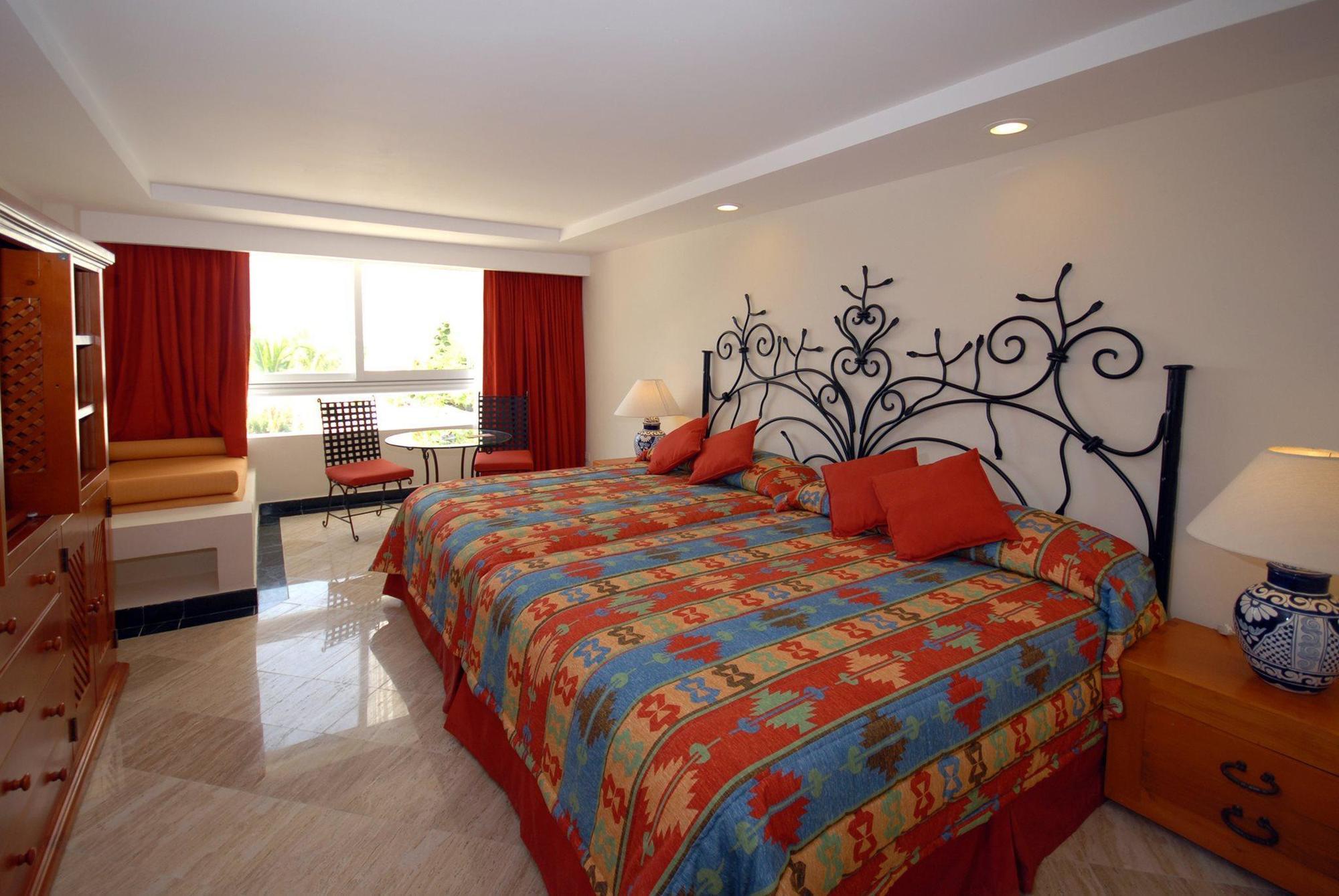 Отель Grand Oasis Palm Канкун Экстерьер фото The photo shows a hotel room with a bright and colorful design. It features two beds with ornate, decorative headboards. The bedding has a vibrant pattern that includes various colors. There are red curtains covering the windows, which allow natural 
