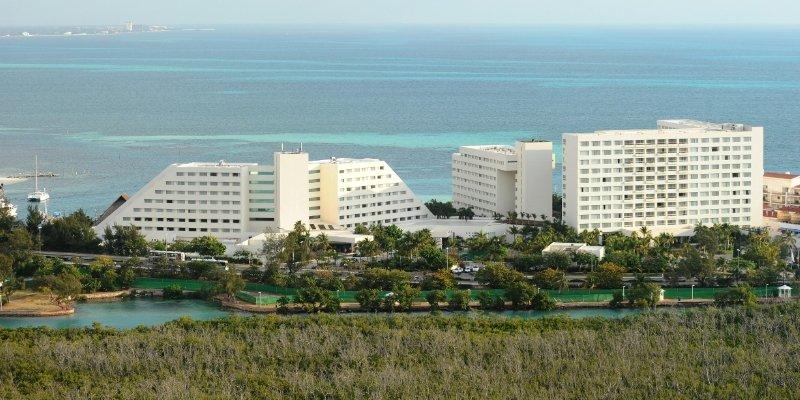 Отель Grand Oasis Palm Канкун Экстерьер фото The photo shows a coastal scene featuring several large hotel buildings. These structures are modern, with a mix of white walls and geometric shapes. They are situated near the ocean, which has clear turquoise waters. In the foreground, there is gree