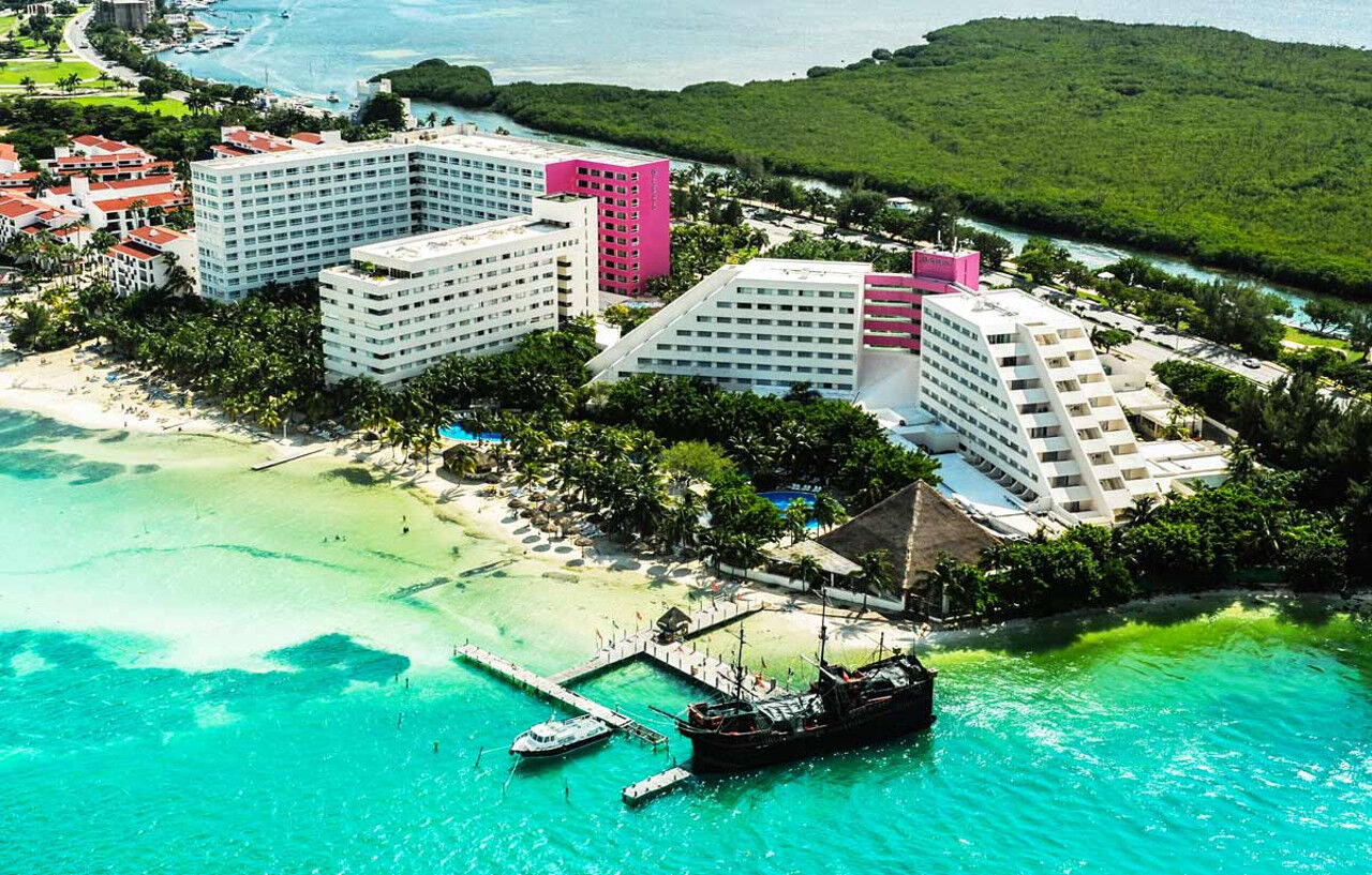 Отель Grand Oasis Palm Канкун Экстерьер фото The photo shows an aerial view of a resort area located near a beach. There are several large buildings, with a mix of white and pink architecture. The buildings are surrounded by lush greenery and palm trees, creating a tropical atmosphere. 

In the